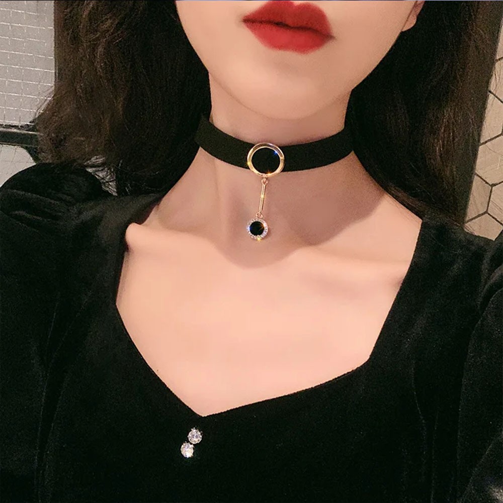 Close no pingente da choker preta, destacando o detalhe em strass e o acabamento dourado, com foco na textura do feltro