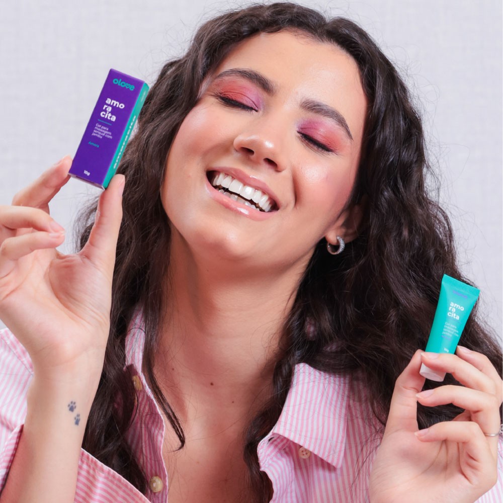 Modelo sorrindo, segurando o Gel Vibrador L�quido Beij�vel Amoracita em sua embalagem e frasco, ilustrando a discri��o e a alegria do uso do produto.
